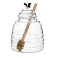 Clear glass beehive-shaped jar with wooden dipper and decorative bee on a white background