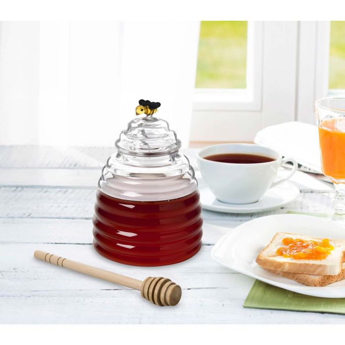 Glass honey dipper with honey, a cup of tea, and toast with jam on a white surface.