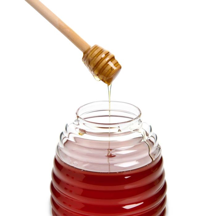 Honey dipper drizzling honey into a glass jar on a white background